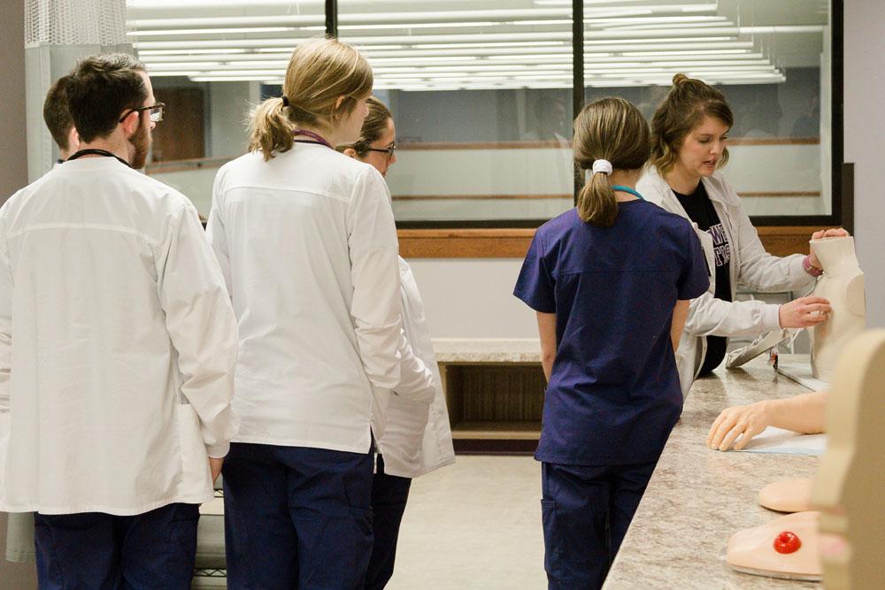 Nursing instructor conducts demonstration on mannequin with nursing students watching