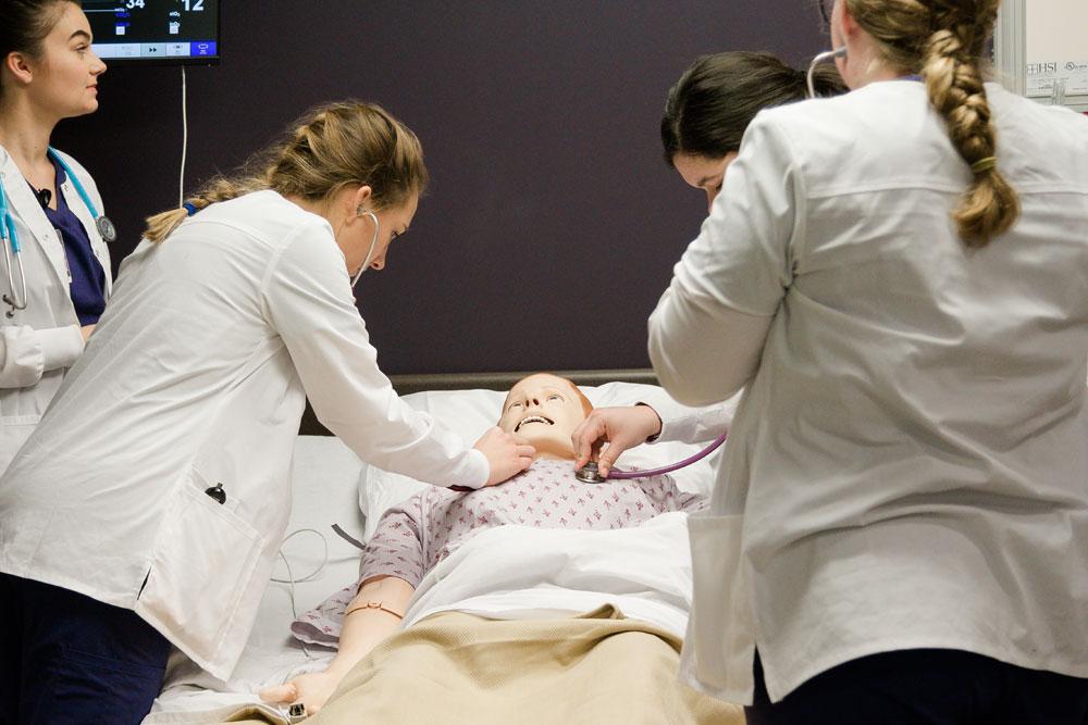 Group of nursing students use stethoscopes to listen to mannequin's heart beat 