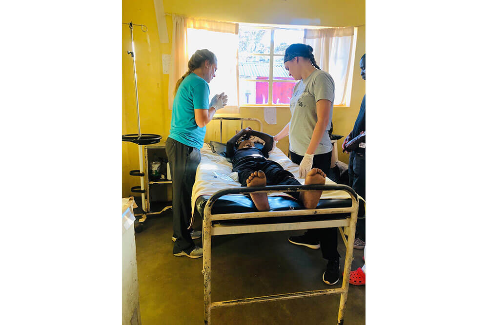 Two female physical therapy students stand over patient lying in bed