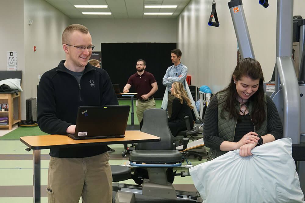 male instructor stands at laptop while female student carries supplies