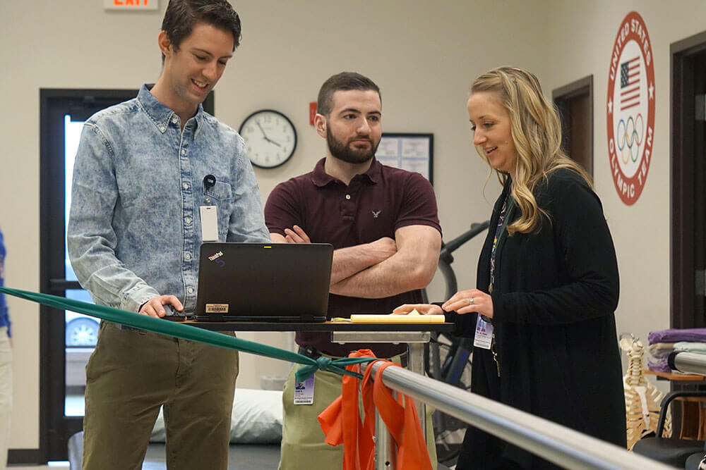 two male students stand behind laptop screen as female instructor provides feedback