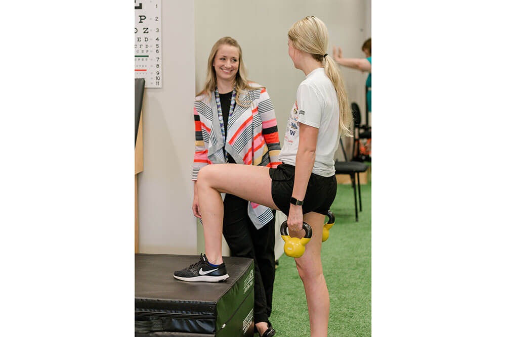 Female professor smiles while watching female student do physical therapy exercises