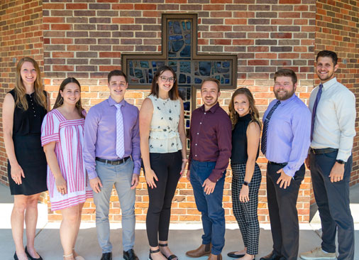 eight admissions counselors pose for picture in front of stained glass cross window