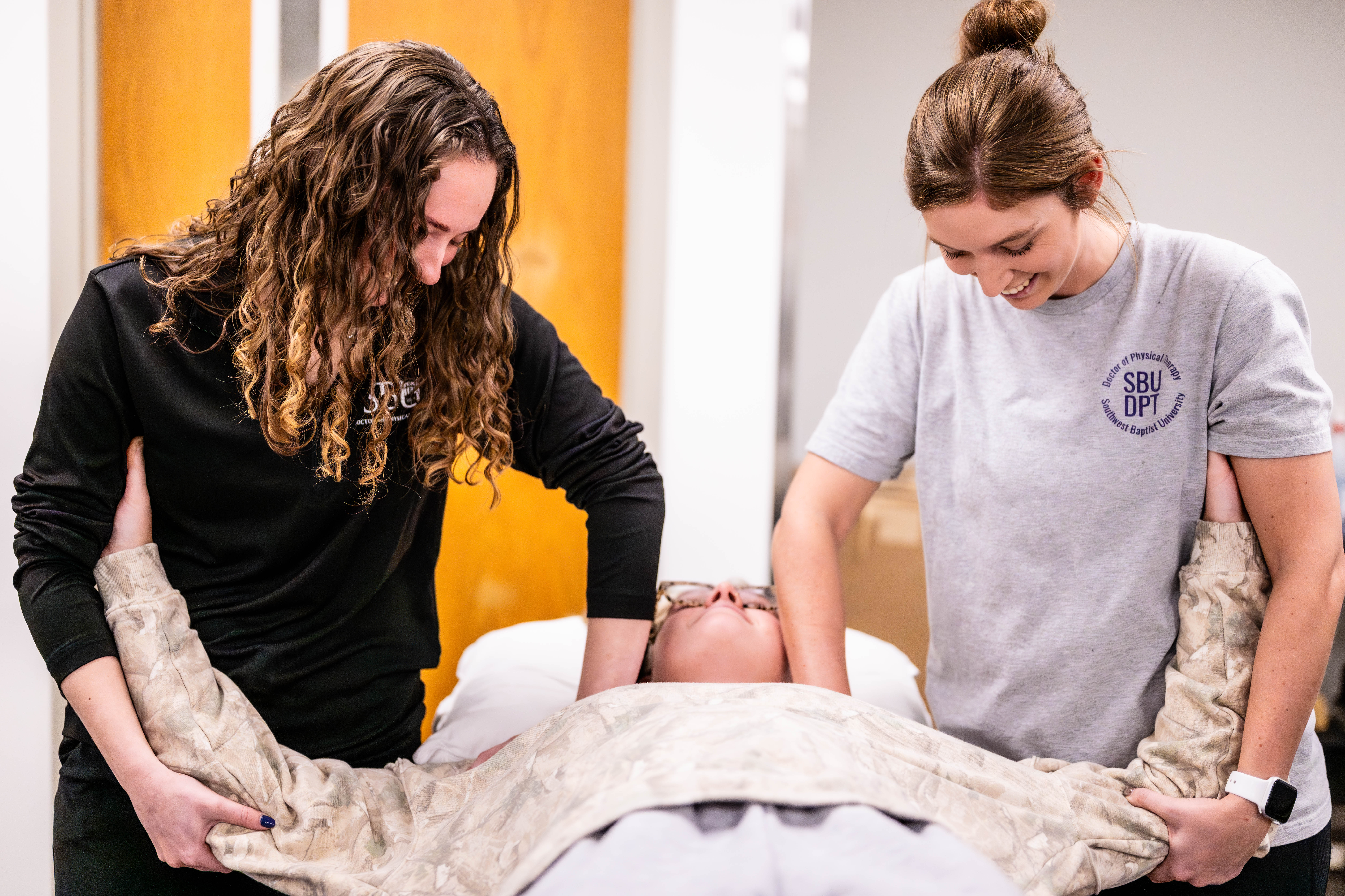 Physical therapy professor demonstrates an exercise on a student laying on lab table while explaining to another student looking on