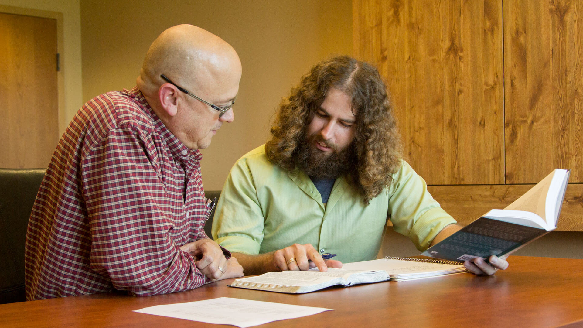 professor and student sit at table looking at Bible and discussing it with each other