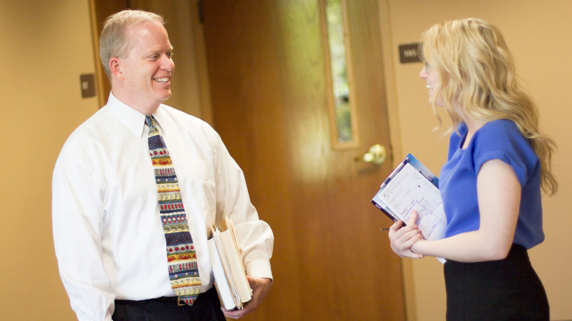 professor holds books and stands in hallway talking to student