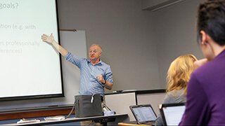Professor points to screen as he lectures in front of students taking notes on laptops