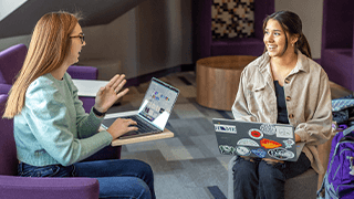 two girls sit on purple chairs in building lobby smiling and talking to each other while working on laptops
