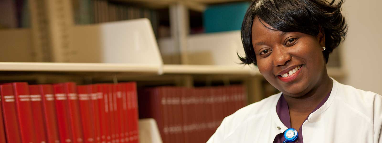 smiling nursing student with stethoscope around neck standing in front of bookshelf