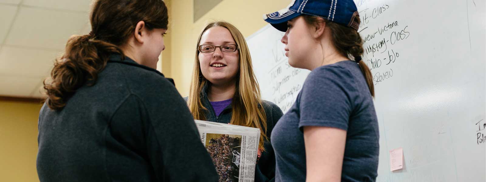 three students standing and talking in front of whiteboard