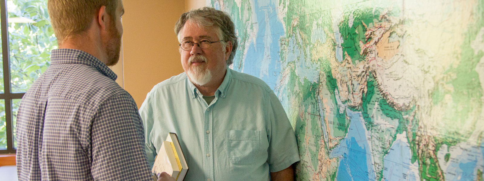 ministry professor holds books and talks to student while standing in front of world map on wall